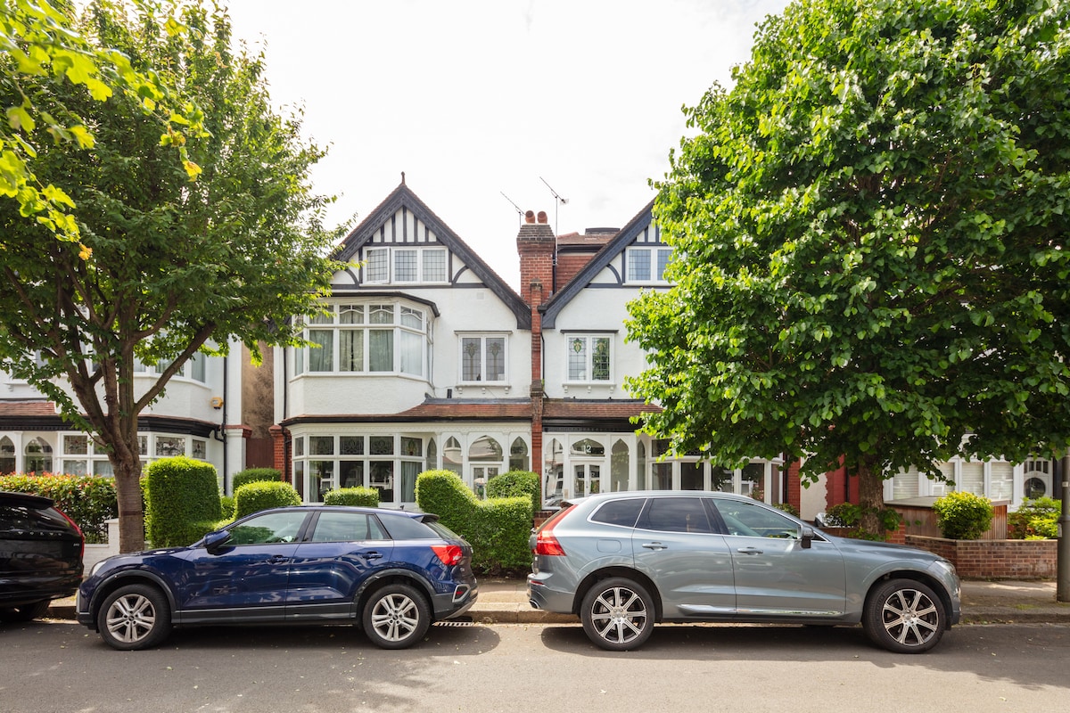 A family home is visible in a quiet residential area, characterized by traditional architecture. Two cars are parked in front of the house, while leafy green trees provide shade along the street. The well-maintained garden features hedges and a neatly landscaped entrance.