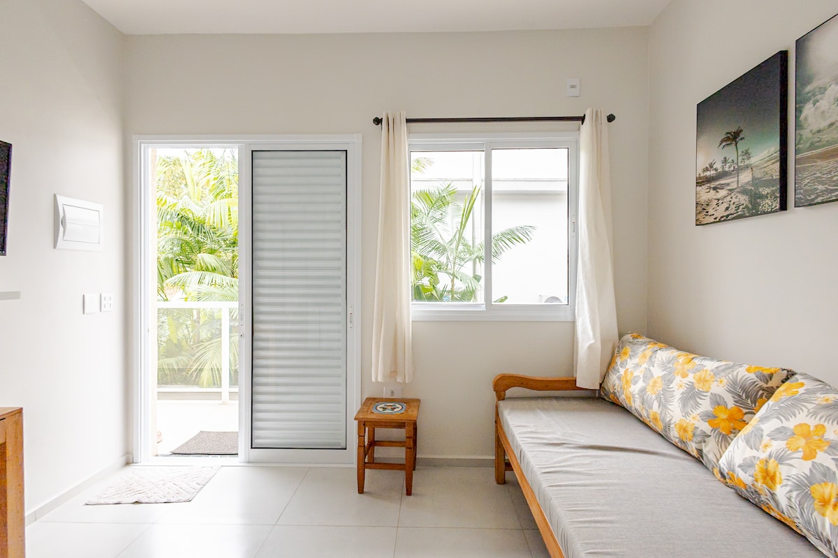 A bright living area features a sofa adorned with floral upholstery, complemented by a wooden side table. Natural light enters through a large window, providing a view of lush greenery outside. A sliding glass door offers access to the balcony.