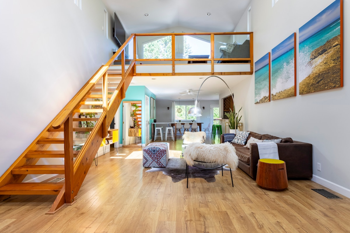 The main living area features high vaulted ceilings and wooden beams, with a staircase leading to the loft. A large brown sofa is centered around a fur throw and round side table, and colorful artwork adorns the wall, complementing the bright and airy ambiance.