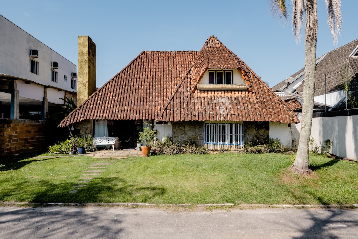 A charming house features a distinctive thatched roof and stone walls, surrounded by well-maintained grass. A pathway leads to the entrance, complemented by potted plants and palm trees that offer a touch of nature to the exterior.