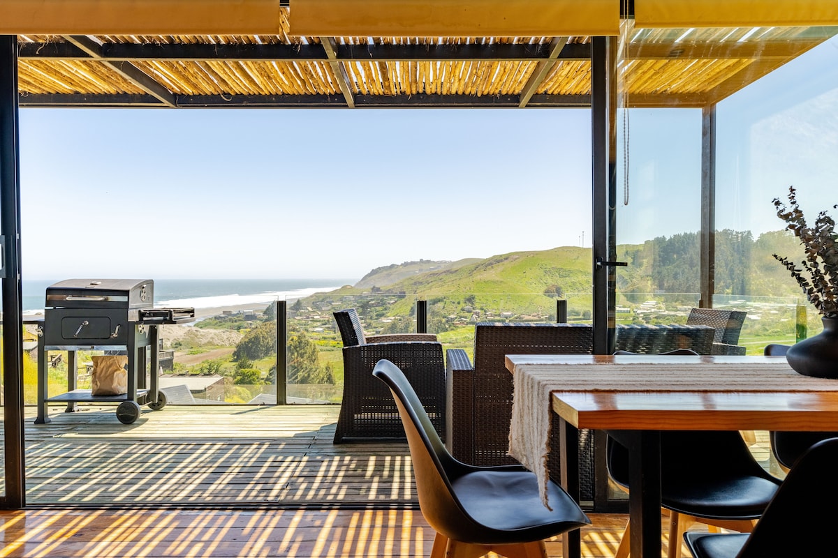 An expansive deck offers a relaxing seating area with wicker chairs positioned around a wooden dining table. A charcoal grill is visible, alongside large glass doors that frame stunning views of the ocean and surrounding hills.