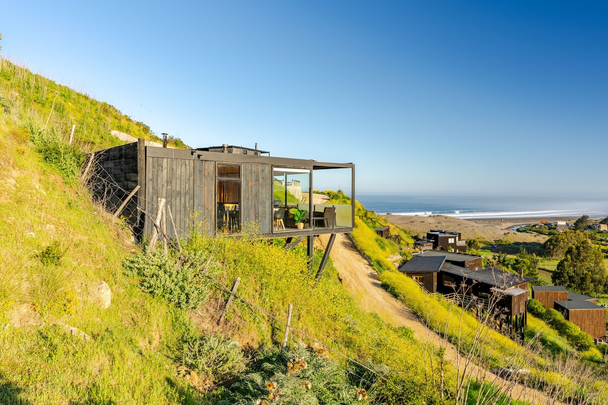 A unique accommodation is presented, featuring large glass windows that provide expansive views of the coastline and surrounding landscape. The structure is nestled in a grassy area, highlighting its integration with nature and showcasing the tranquility of the environment.
