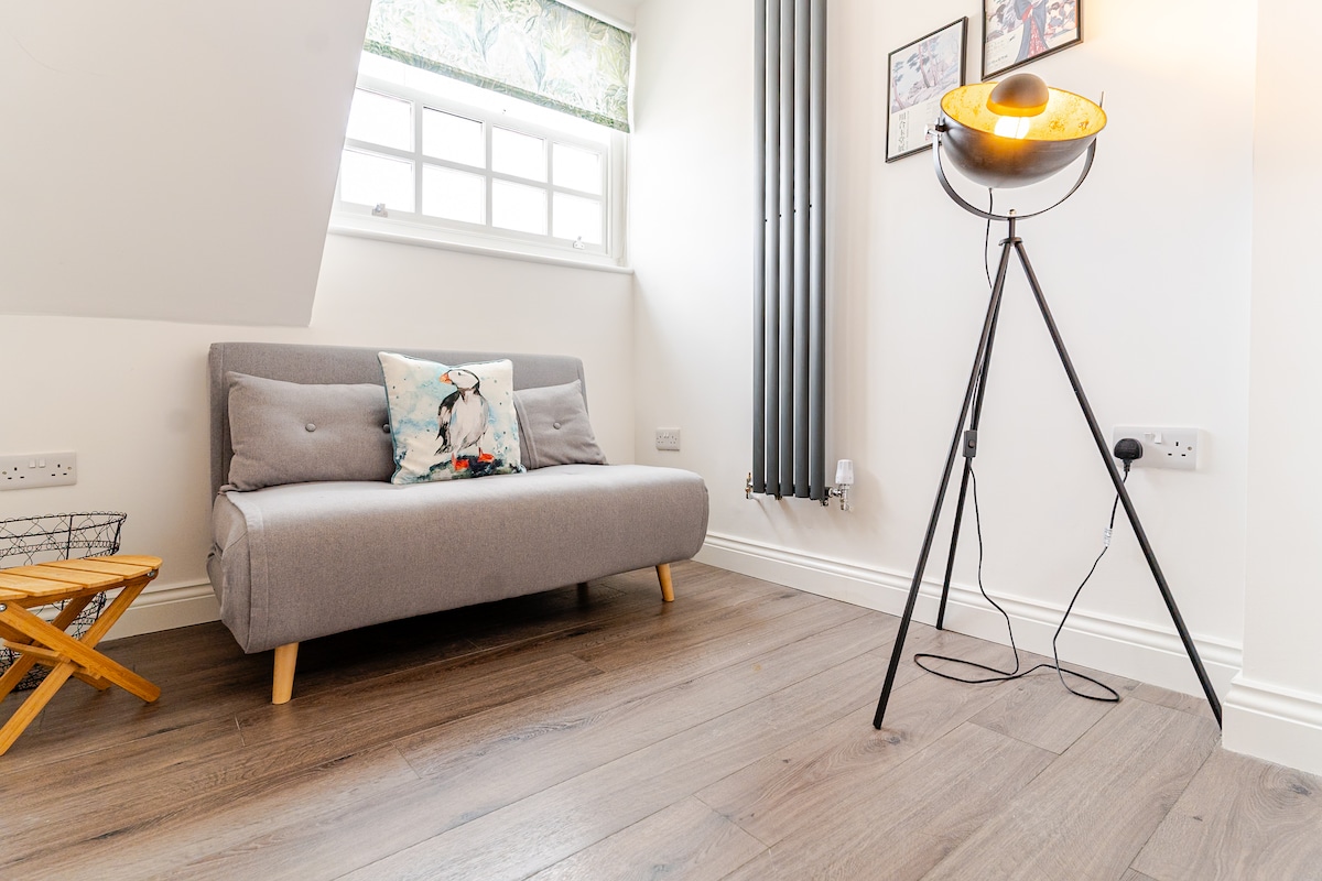 A modern seating area is featured with a grey sofa positioned against a wall. A stylish floor lamp stands nearby, providing soft lighting to the space. Natural light filters in through a window, with a decorative cushion adding a touch of color.