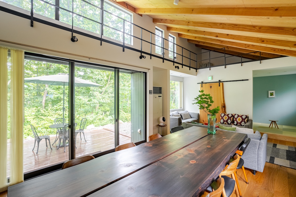 A spacious living and dining area features a large wooden table surrounded by chairs. Floor-to-ceiling windows provide abundant natural light and views of the greenery outside. A cozy seating area with a sofa is visible, contributing to a warm ambiance within the space.