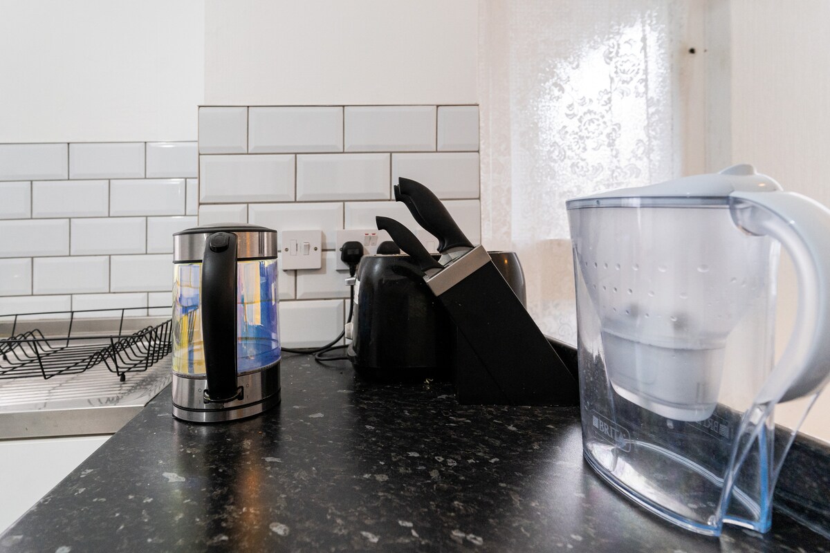 A compact kitchen counter is shown featuring a kettle, a coffee maker, and a water pitcher. Clean white tiles create a bright background, while a drying rack and natural light from a window add to the inviting setting.