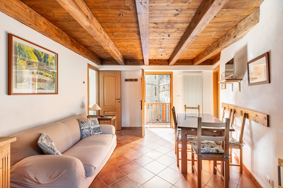 A well-lit living area features a beige sofa and a wooden dining table surrounded by four chairs. Exposed wooden beams run across the ceiling. A door leads to the terrace, with natural light illuminating the space. Artworks adorn the walls, enhancing the ambiance.