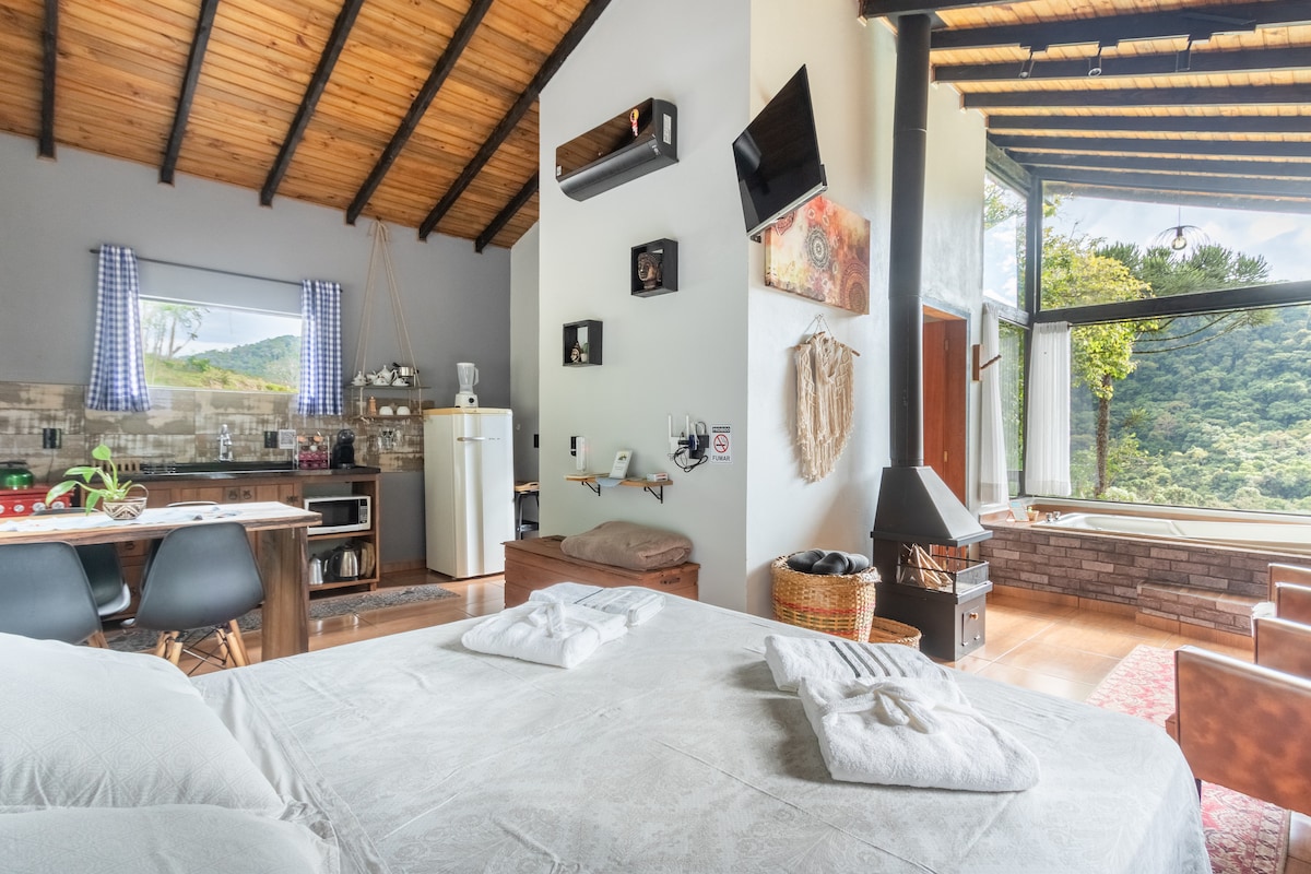 A cozy interior showcases a spacious bed with neatly arranged towels and a view of the surrounding greenery through large windows. A dining area with black chairs is visible beside a well-equipped kitchen, while wooden beams and an accent wall add warmth to the space.