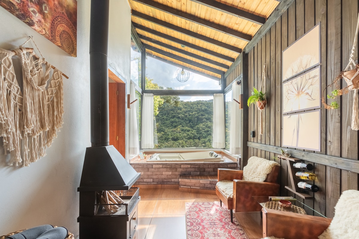 A cozy sitting area features two leather armchairs and a decorative rug. Large windows provide a view of the surrounding greenery. A wood-burning stove stands in one corner, and a small wine rack is also present, adding to the inviting atmosphere of the space.
