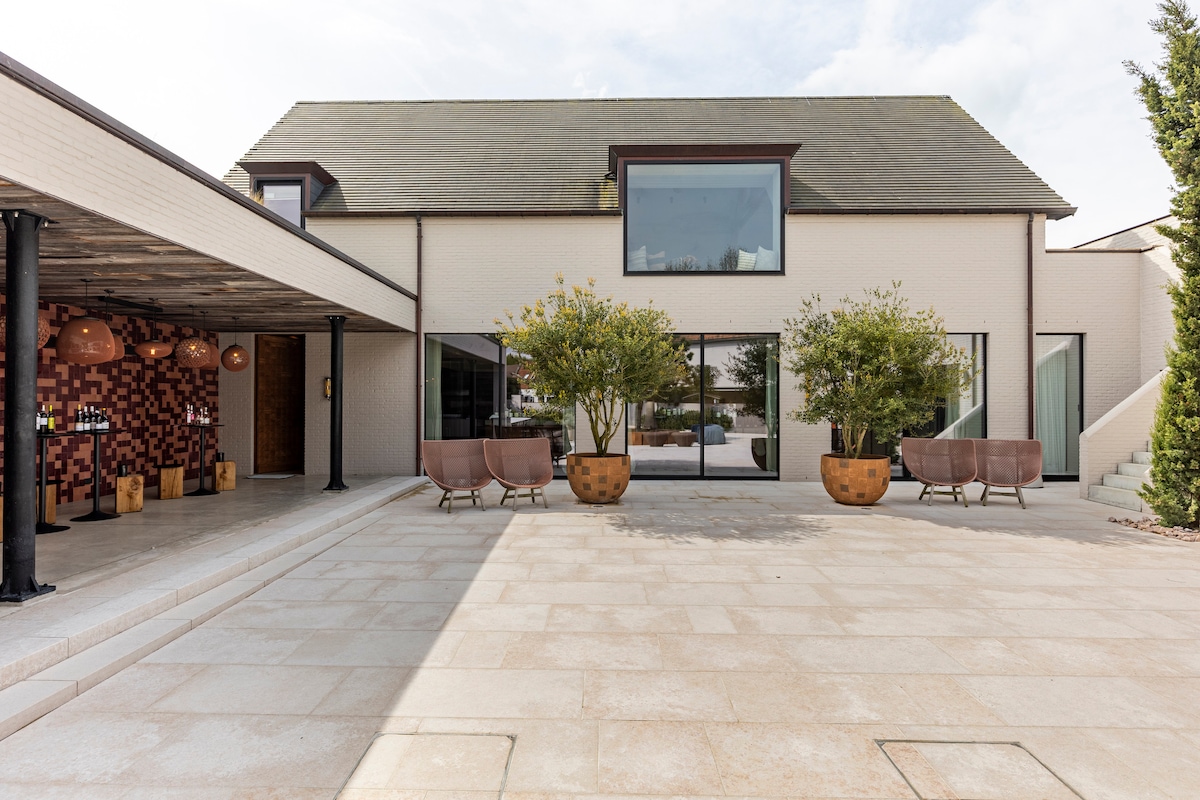 A spacious outdoor area is highlighted by a modern building with large windows. Potted trees add greenery, while distinctive seating arrangements create inviting pockets for relaxation. The stone patio's smooth surface complements the contemporary architecture, enhancing the overall aesthetic.