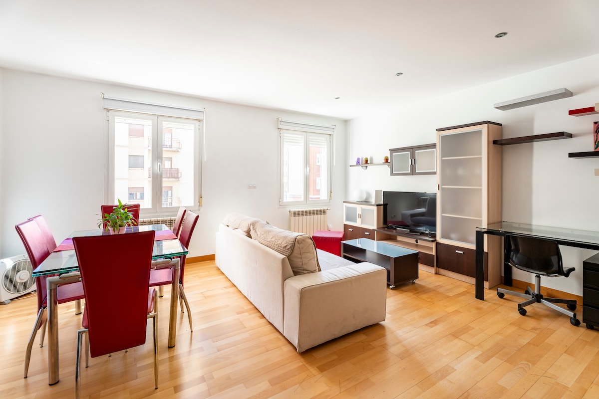 A bright living space featuring a comfortable sofa, a glass dining table surrounded by red chairs, and a wall-mounted entertainment center. Large windows allow natural light to fill the room, while warm wooden flooring complements the modern furniture arrangement.