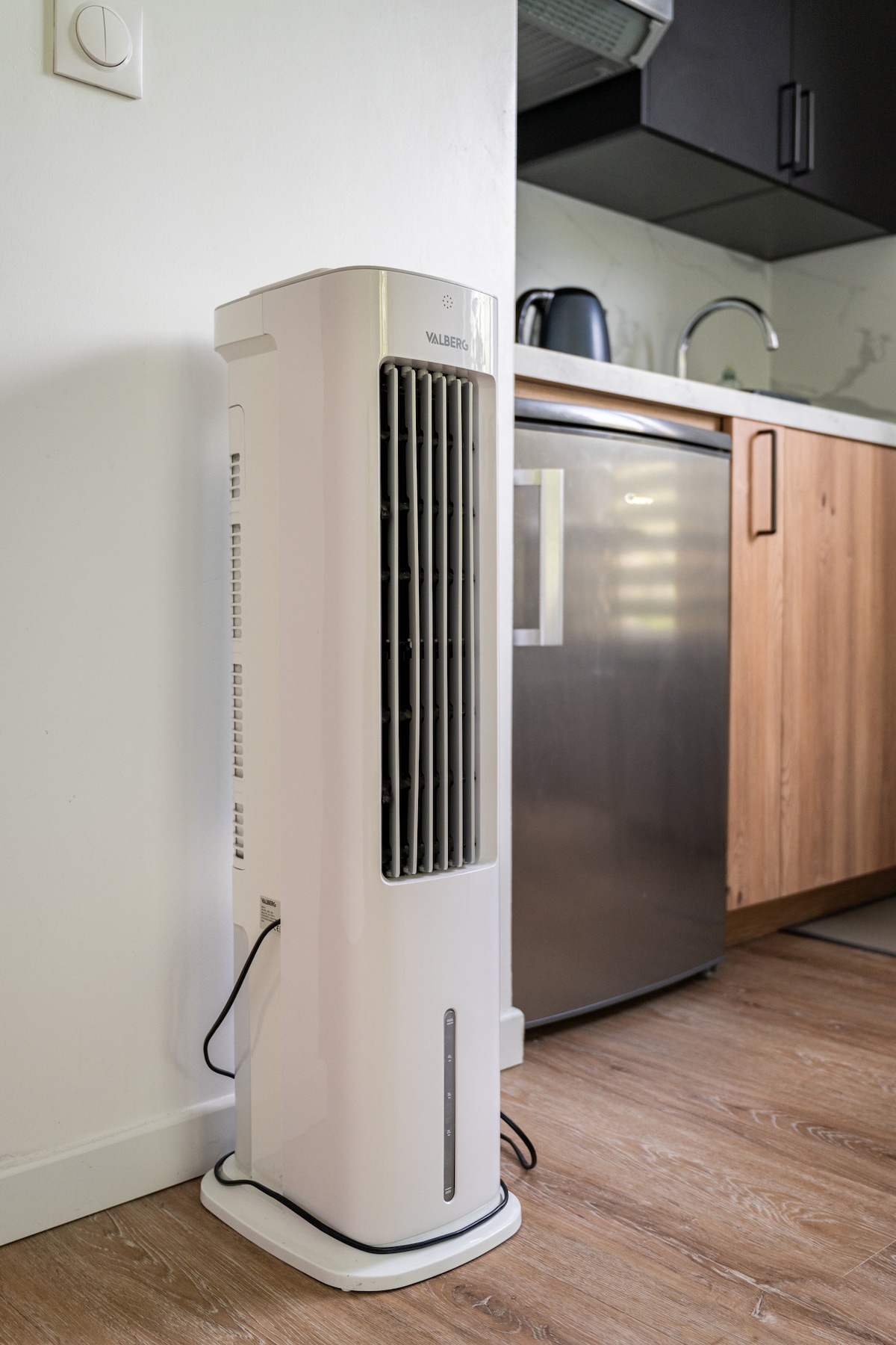 A modern cooling fan is positioned beside the kitchen area, featuring a sleek white design with vertical slats. Nearby, the stainless steel refrigerator and wooden cabinetry create a functional kitchen space with a harmonious blend of materials.