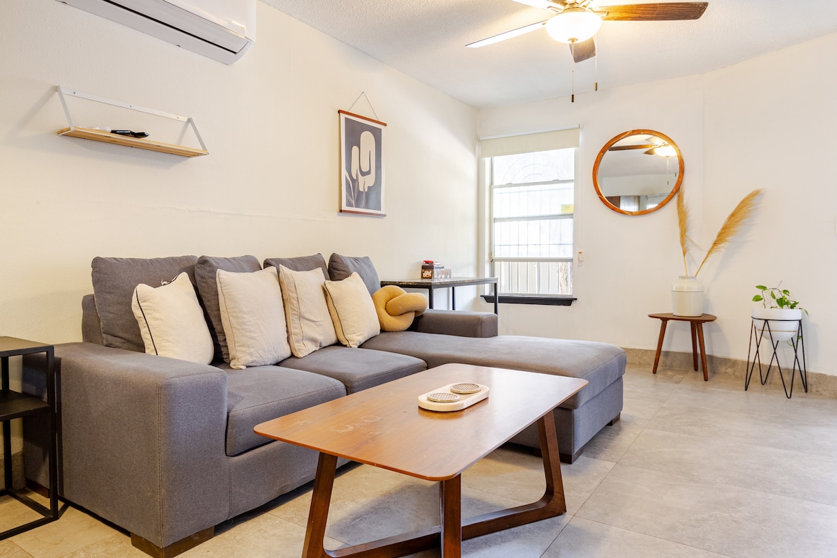 A gray sectional sofa with decorative pillows is positioned in the living area. A wooden coffee table is displayed in front, accompanied by a small round mirror and a shelf on the wall. A floor plant and a light-colored chair add to the inviting space.