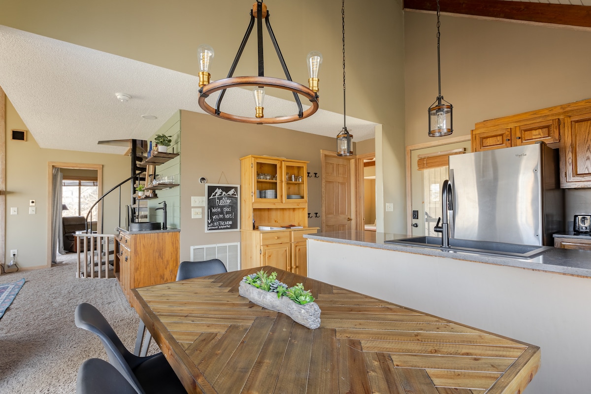 A dining area features a rustic wooden table surrounded by black chairs. Above, a modern chandelier with hanging bulbs provides light. The kitchen is visible in the background, highlighting wooden cabinetry and stainless steel appliances. Natural light enters through nearby windows.