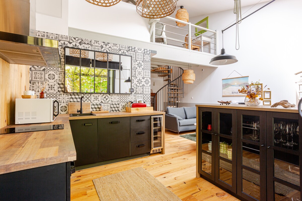 An open kitchen and living area feature modern appliances and stylish cabinetry. A decorative backsplash adds visual interest. A dining area is highlighted by a wooden table, while a cozy seating arrangement is positioned nearby. Natural light filters through large windows, enhancing the space's inviting atmosphere.