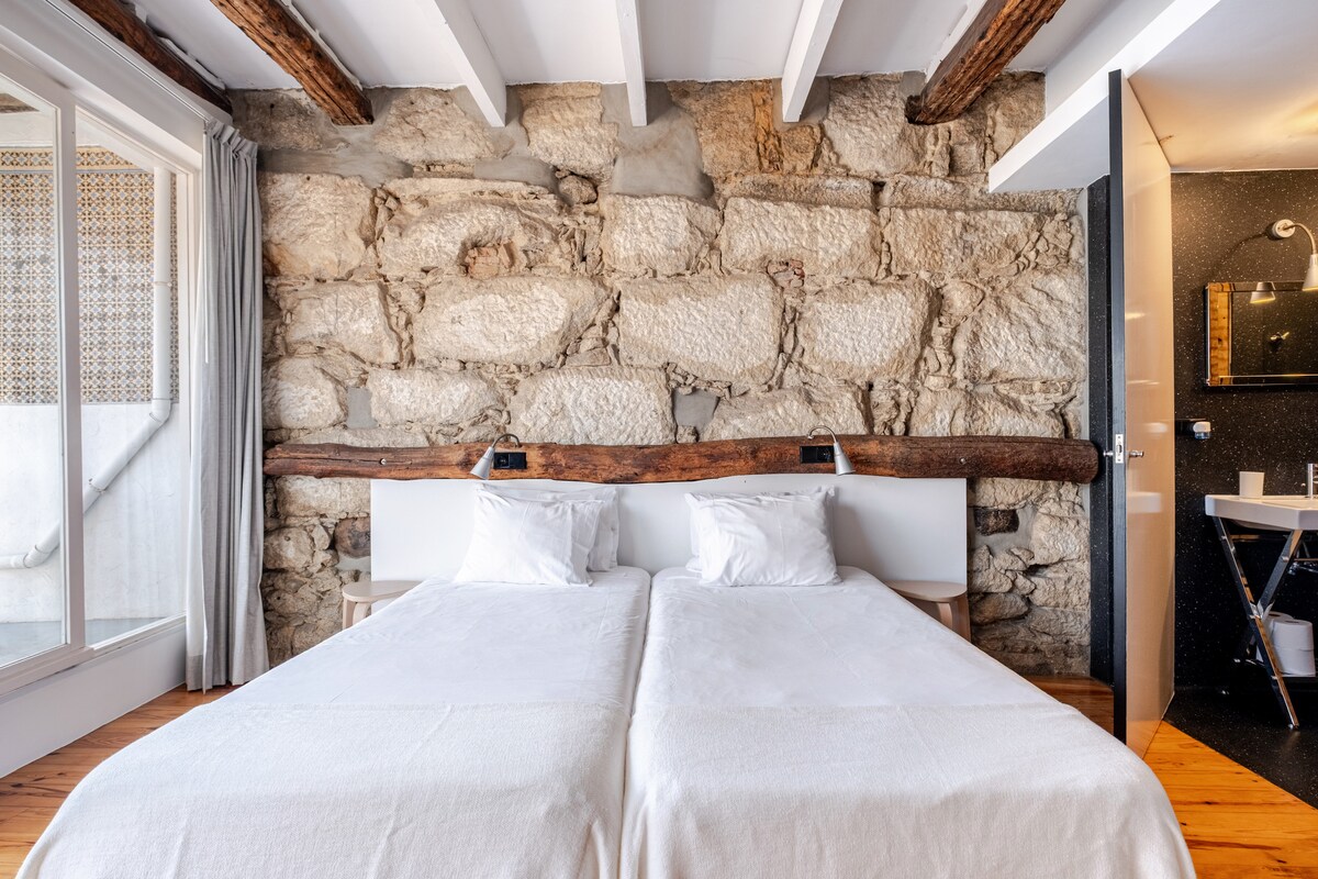 A bedroom features two spacious beds on a warm wooden floor, set against a rustic stone wall. The natural texture of the wall complements the white bedding. A minimalistic ensuite bathroom is visible through an open door, adding a touch of contemporary design.