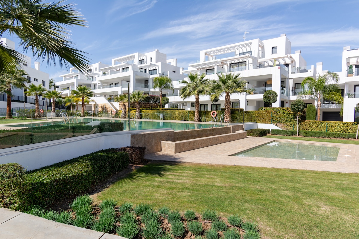 The image showcases a landscaped outdoor area featuring manicured lawns and palm trees. Several pools are visible, with a shallow area and a larger pool that reflects the surrounding architecture. White modern buildings are seen in the background against a clear blue sky.