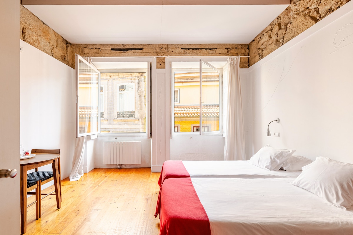 A spacious room features two beds adorned with white linens, complemented by a red blanket. Large windows open to allow natural light, revealing views of the historic architecture outside. A simple wooden table is positioned nearby, enhancing the room's functionality.