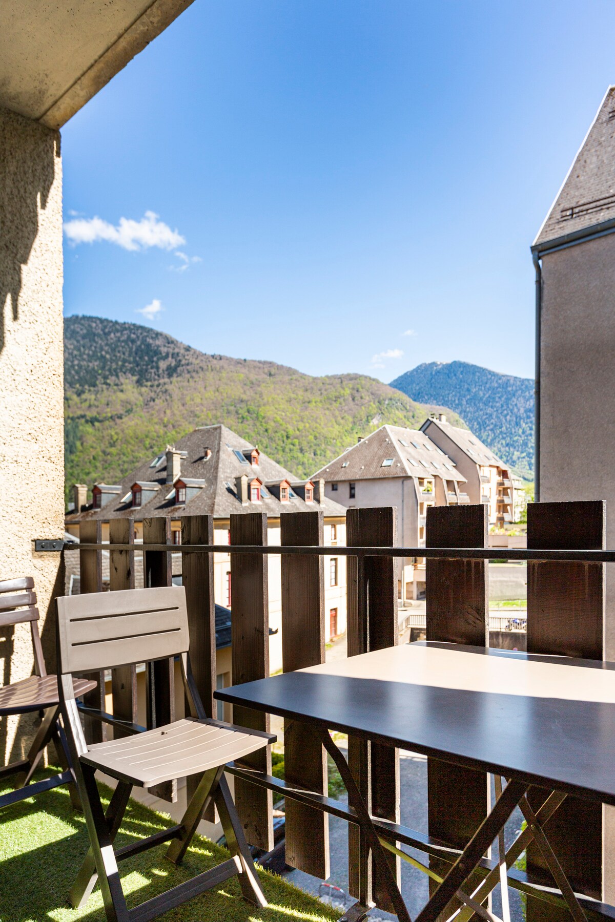 A balcony view is presented, featuring wooden railings and a small table. Two chairs are positioned for seating. In the background, mountains rise with greenery, bathed in sunlight, creating a serene atmosphere. Clear blue skies enhance the overall landscape.