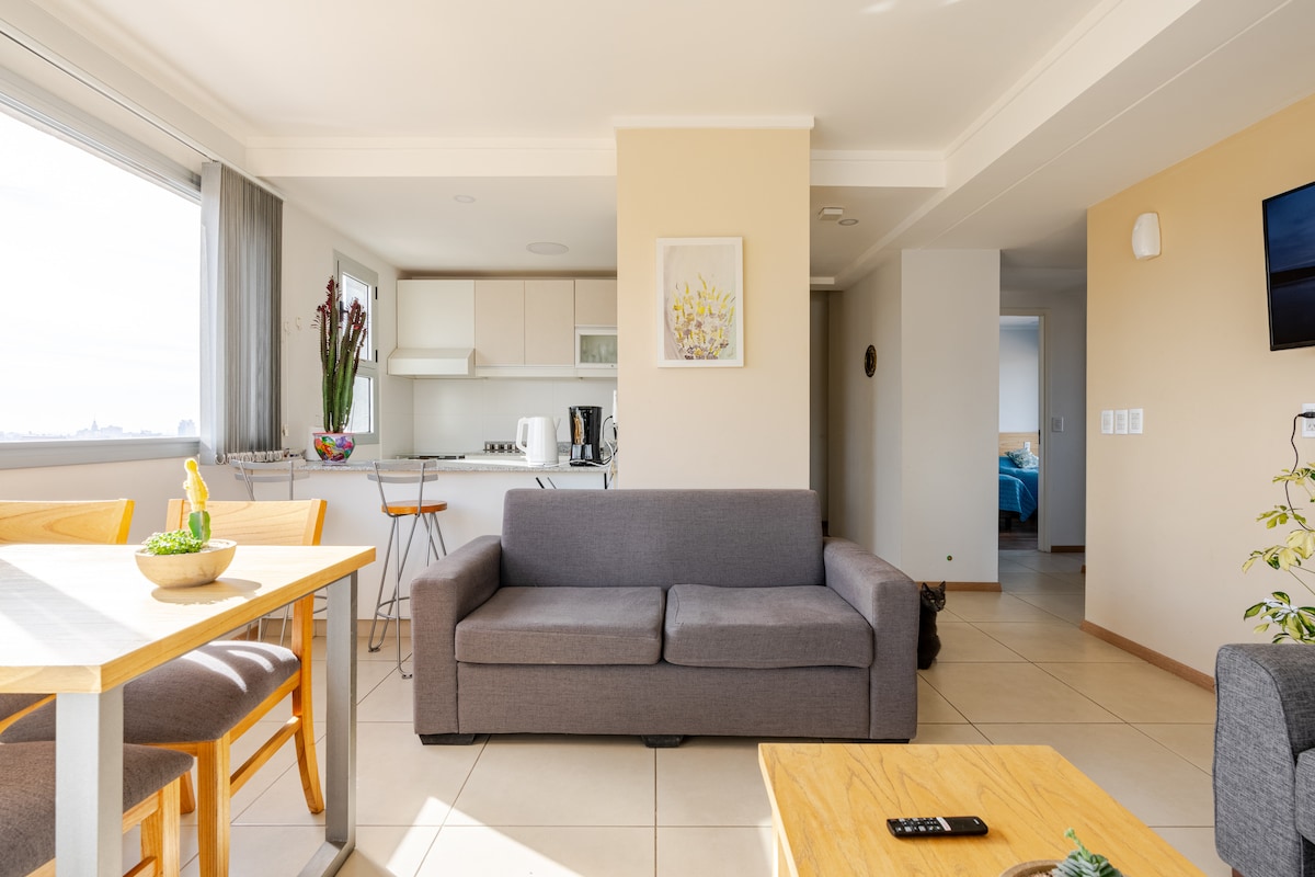 A modern living space features a comfortable gray sofa facing a wooden coffee table. A dining area with four chairs is positioned next to a bright window, allowing natural light to fill the room. The open kitchen is visible in the background, equipped with essential appliances.