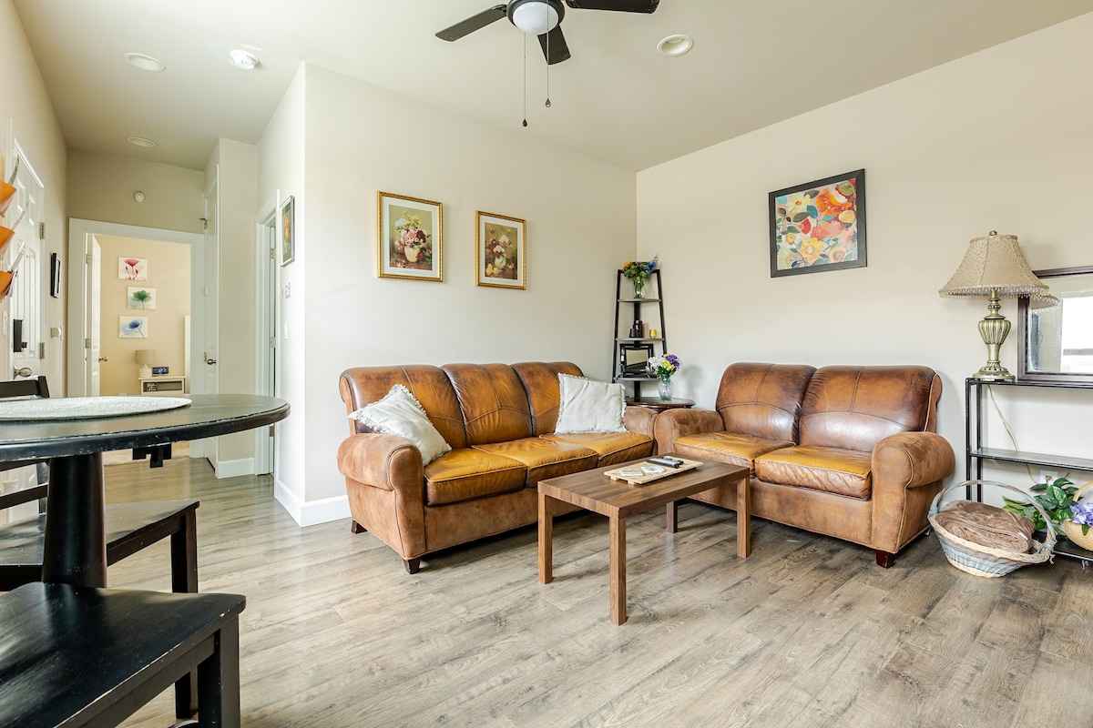 A comfortable living area is presented with two brown leather sofas and a wooden coffee table. Light gray walls provide a neutral backdrop, while framed artwork adds color. A ceiling fan is visible above, and natural light enters through a nearby door.
