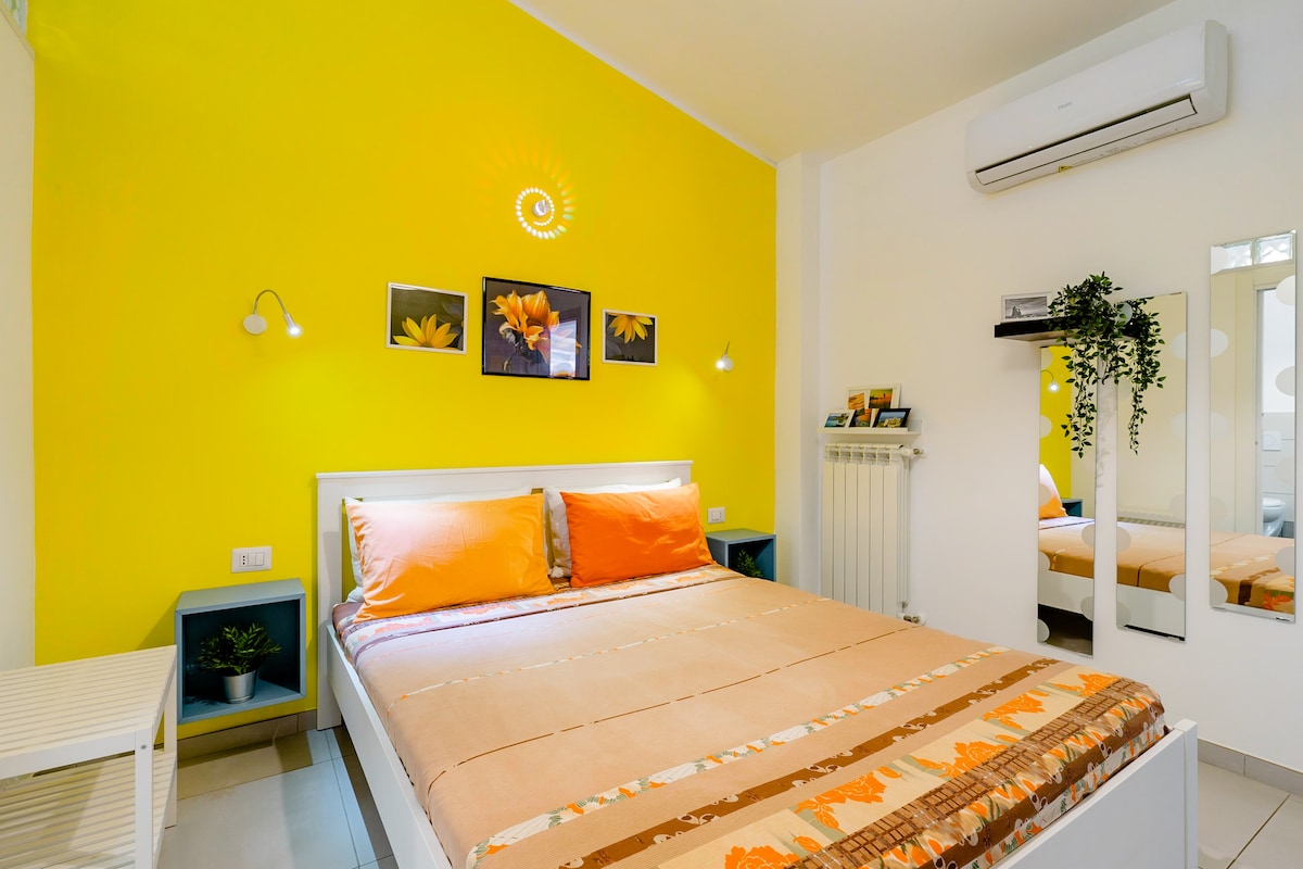 The room features a spacious bed with orange and patterned blankets, complemented by bright yellow walls. Two framed flower prints are displayed above the bed. Soft lighting is provided by wall sconces, and a full-length mirror is mounted on the wall opposite the bed.