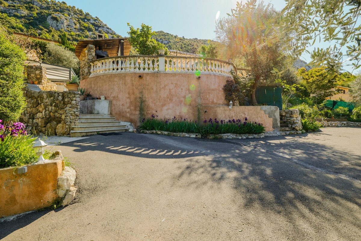 A serene exterior view of the villa is presented, featuring a circular terrace with a decorative railing. Lush greenery surrounds the entrance, and the driveway is lined with blooming flowers. Sunlight filters through the trees, creating a calming atmosphere in the landscape.