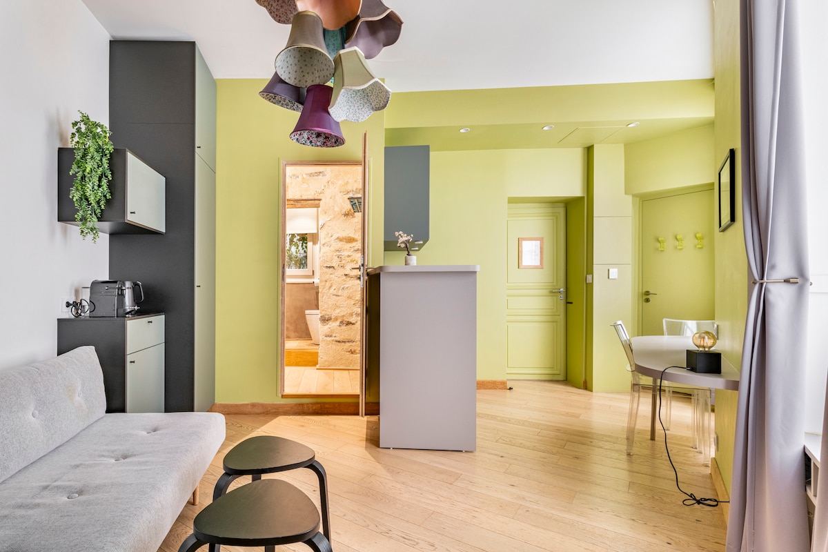A modern living space features a light-colored sofa and two small dark tables. The walls are painted in soft green, complemented by a unique multi-colored pendant light. A kitchenette is visible in the corner, while a doorway leads to a well-lit area beyond.