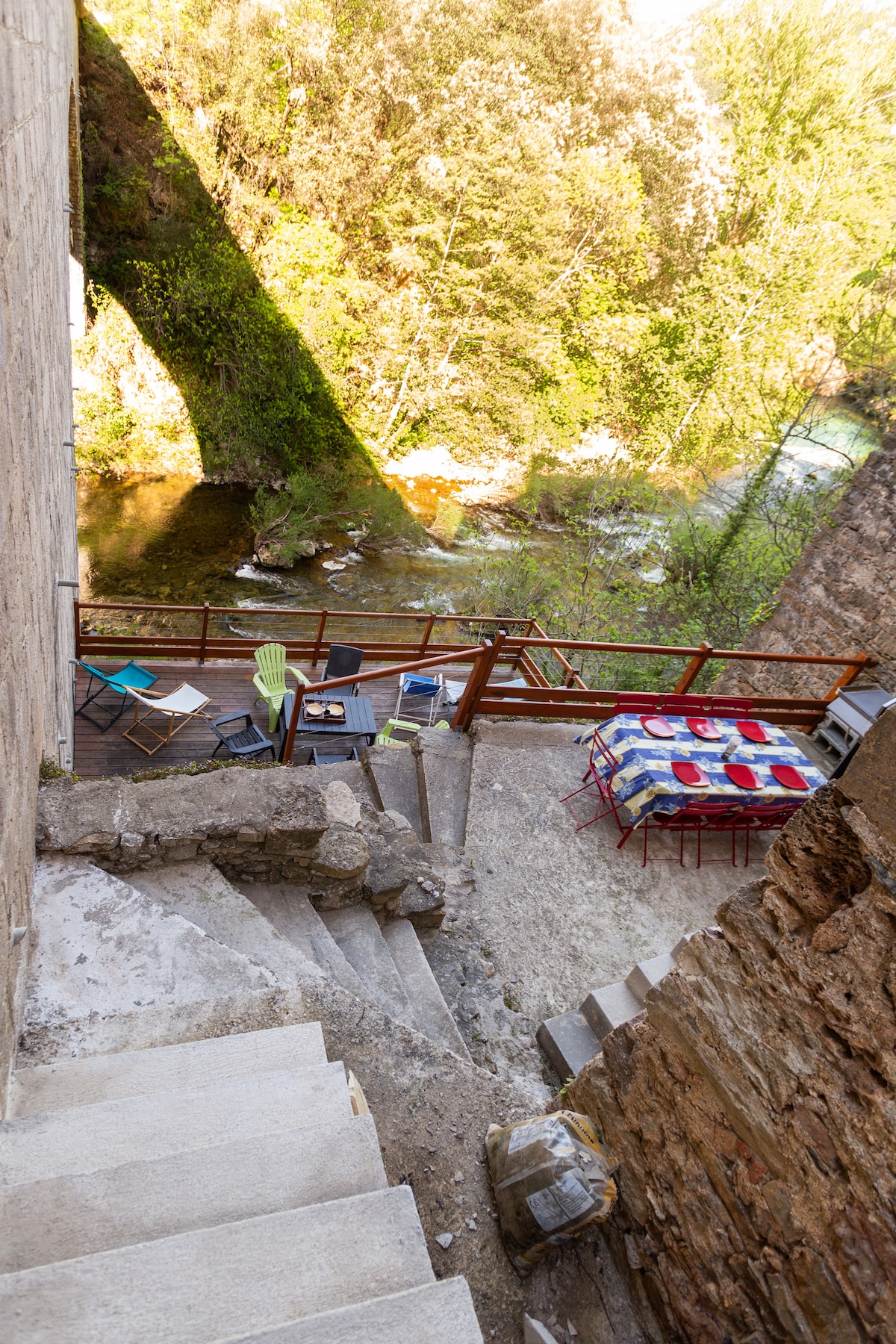 A serene terrace area is visible, featuring a dining table set with vibrant tablecloth, surrounded by a mix of chairs and loungers. Stairs lead down to a natural setting with a river and greenery, creating an inviting space for relaxation and outdoor meals.