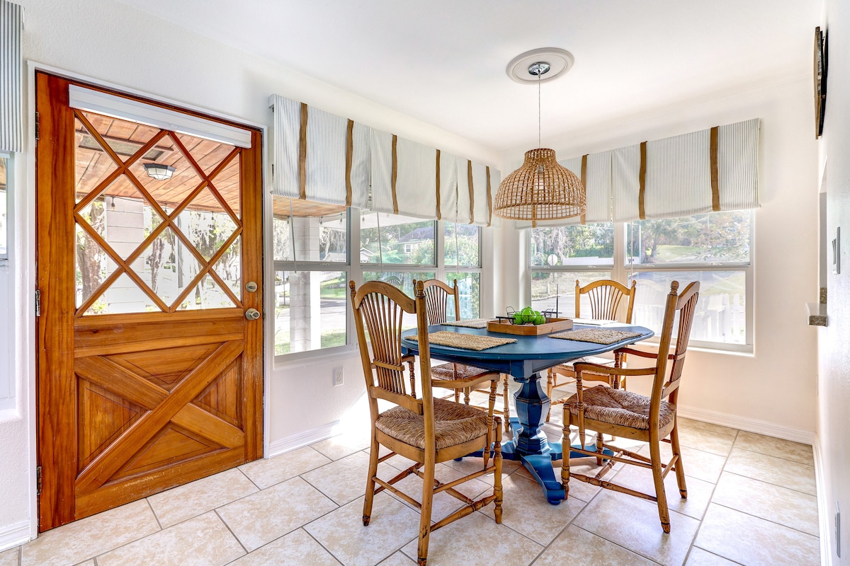 A cozy dining area is illuminated by natural light from windows on three sides. A blue table is surrounded by four wooden chairs, and a woven pendant light hangs above. Decorative elements include a bowl of fruit on the table, enhancing the inviting atmosphere.