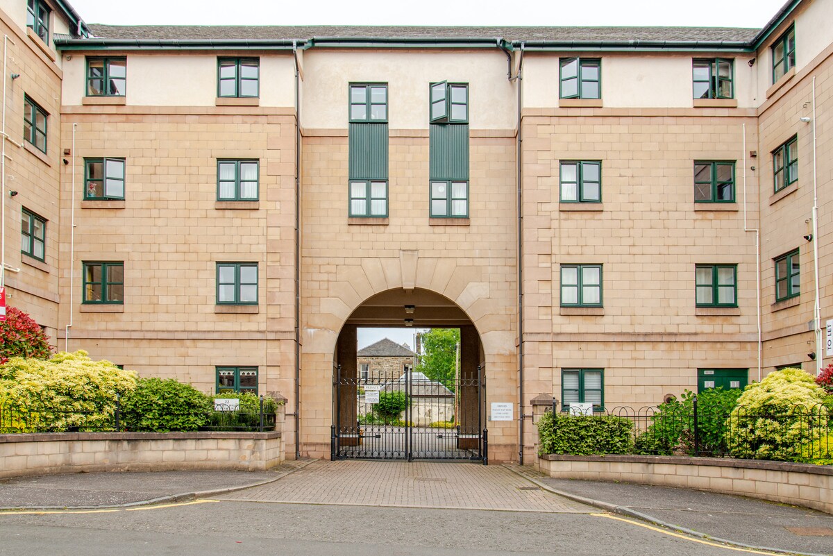 The exterior view of the apartment building reveals a modern structure with a stone façade and multiple windows. An arched entranceway is highlighted, providing access to the interior courtyard. Lush greenery surrounds the perimeter, offering a welcoming sight for guests.