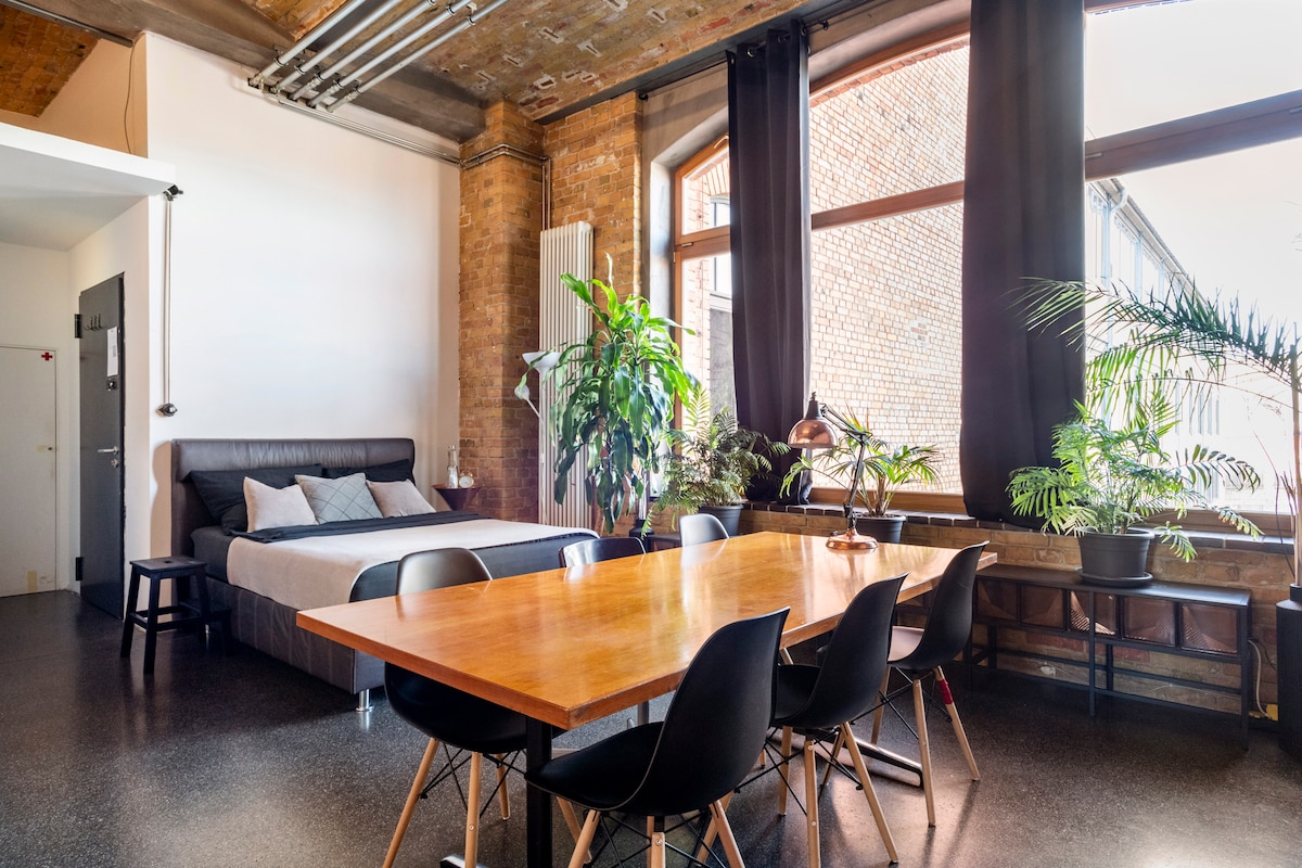 An open living space features a spacious bed against a brick wall, complemented by tall windows adorned with dark curtains. A large wooden dining table with six chairs occupies the center, while lush green plants add a touch of nature to the room.