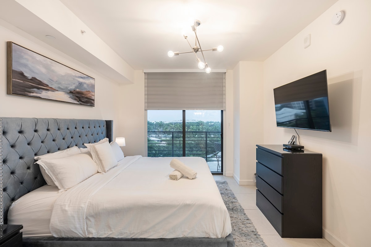 A spacious bedroom features a tufted bed with crisp linens, complemented by modern decor. A large flat-screen TV is mounted on the wall, and a stylish dresser provides additional storage. Natural light fills the room through a partially closed window shade, offering a view of the outdoors.