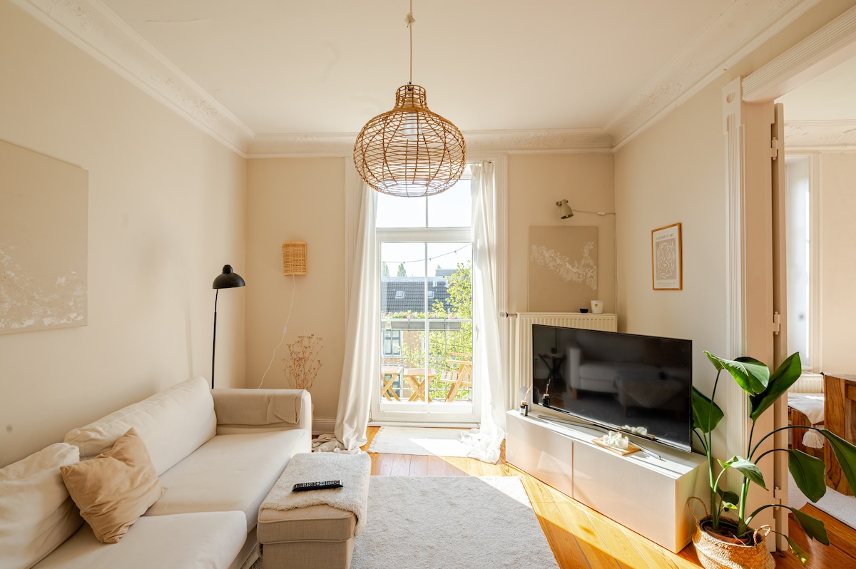 A light-filled living room features a comfortable white sofa with soft pillows. A large window with sheer curtains provides direct access to the balcony, while natural light illuminates the room. A modern television sits on a low console, accompanied by a potted plant and a floor lamp.