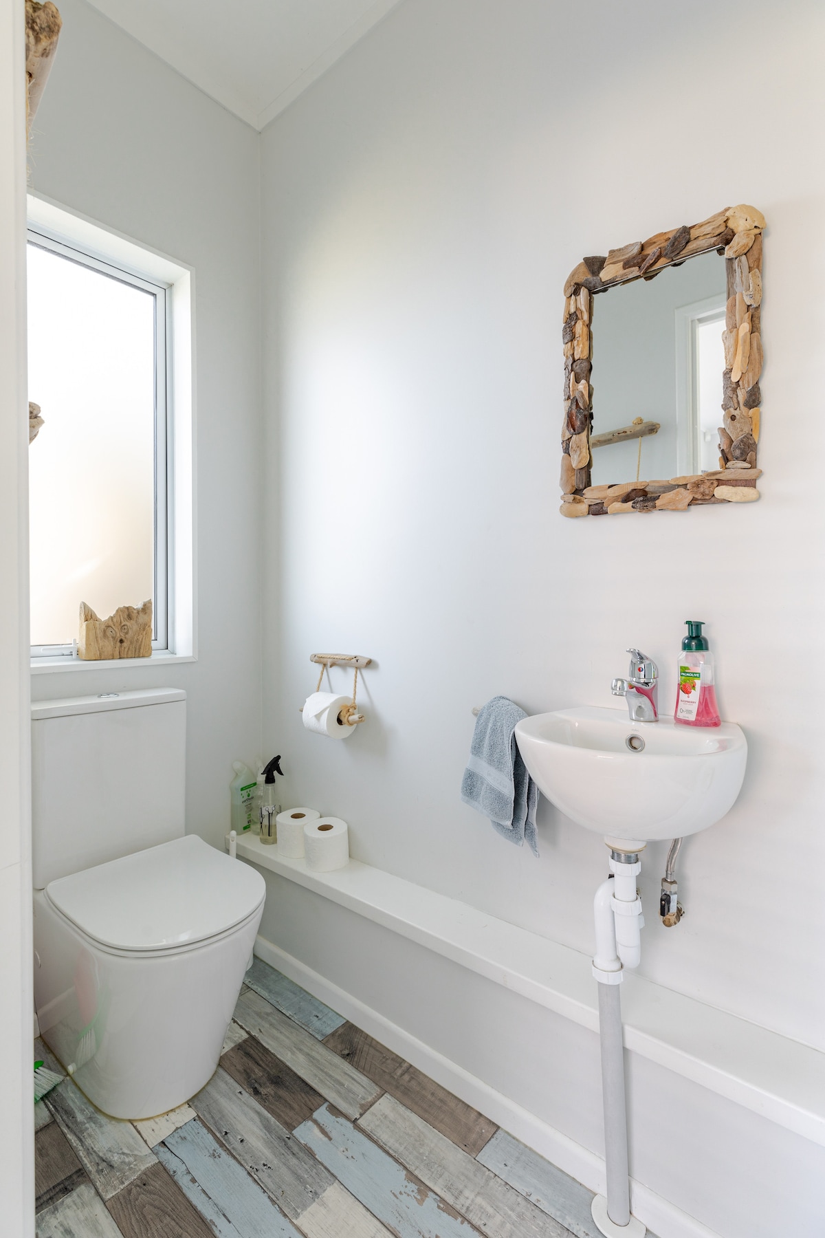 A compact bathroom is presented, featuring a toilet and a small sink mounted on a wall. Natural light is let in through a nearby window. A mirror framed with natural materials hangs above the sink, while a towel and bathroom essentials are neatly arranged nearby.