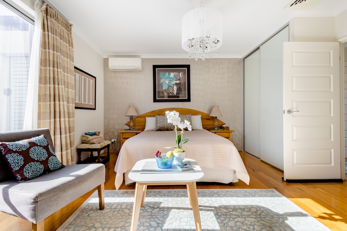 A well-appointed studio bedroom features a queen-sized bed with a light-colored bedspread, complemented by decorative pillows. Natural light fills the room through large windows adorned with elegant curtains. A cozy seating area with a chair is positioned next to a small table, offering a welcoming touch.