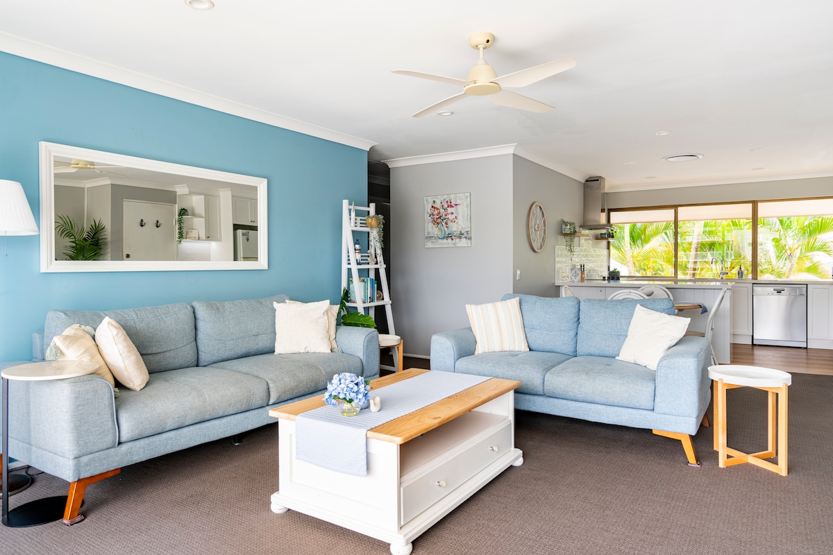 A spacious living area features two light blue couches, complemented by a small white side table and a wooden coffee table with decorative flowers. A ceiling fan provides cooling, while large windows invite natural light and showcase views of greenery beyond.
