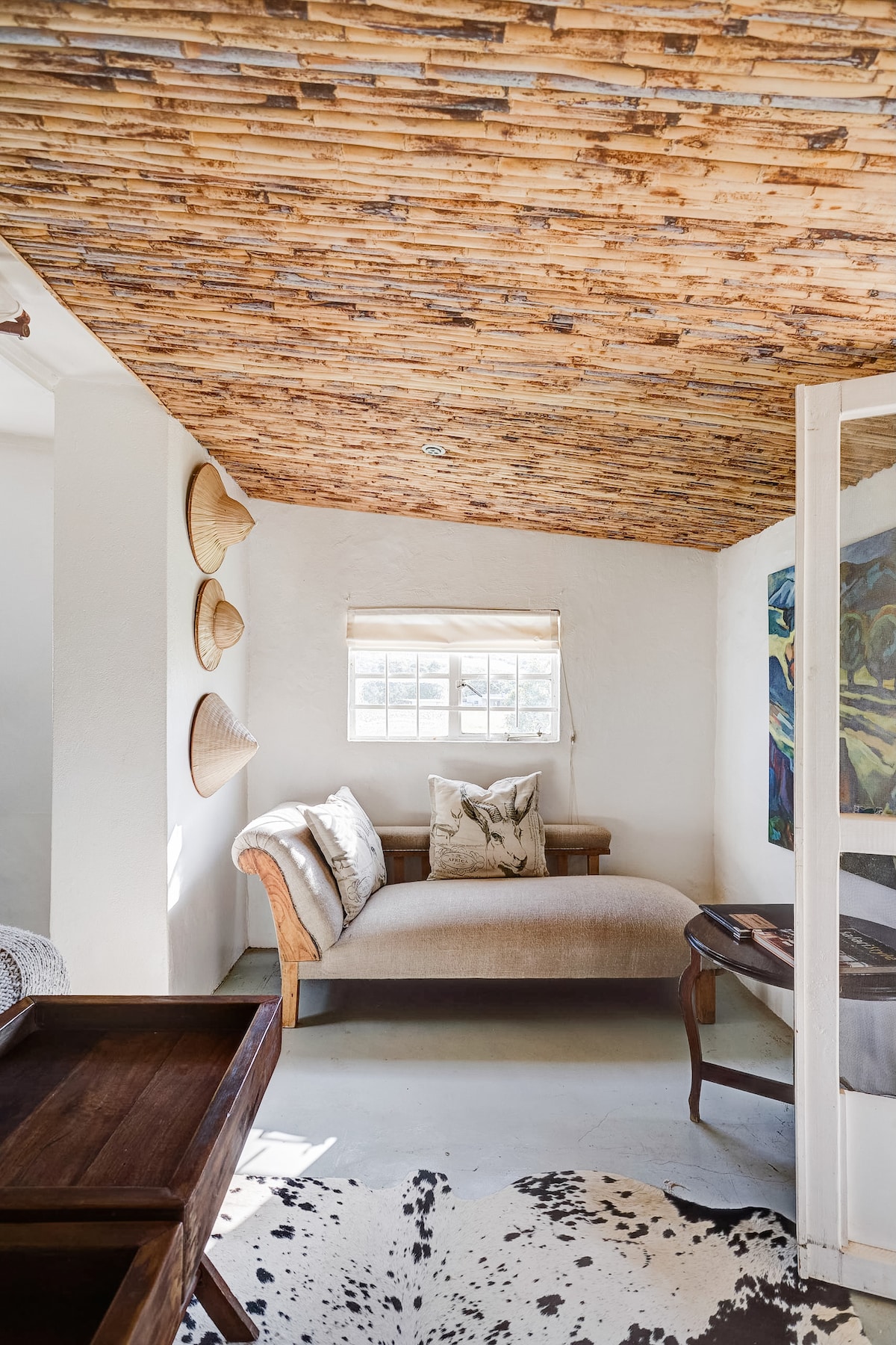 A cozy corner of the cottage features a comfortable chaise lounge adorned with pillows. Natural light enters through a small window dressed with a simple blind. Traditional woven hats are displayed on the wall, complemented by a piece of colorful artwork nearby.