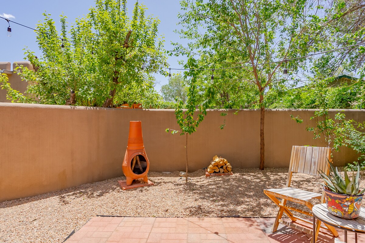 A private, enclosed yard is framed by natural greenery and a beige wall. A clay chiminea is positioned near a stack of firewood. Seating is provided by a wooden chair and a small round table adorned with a potted plant.