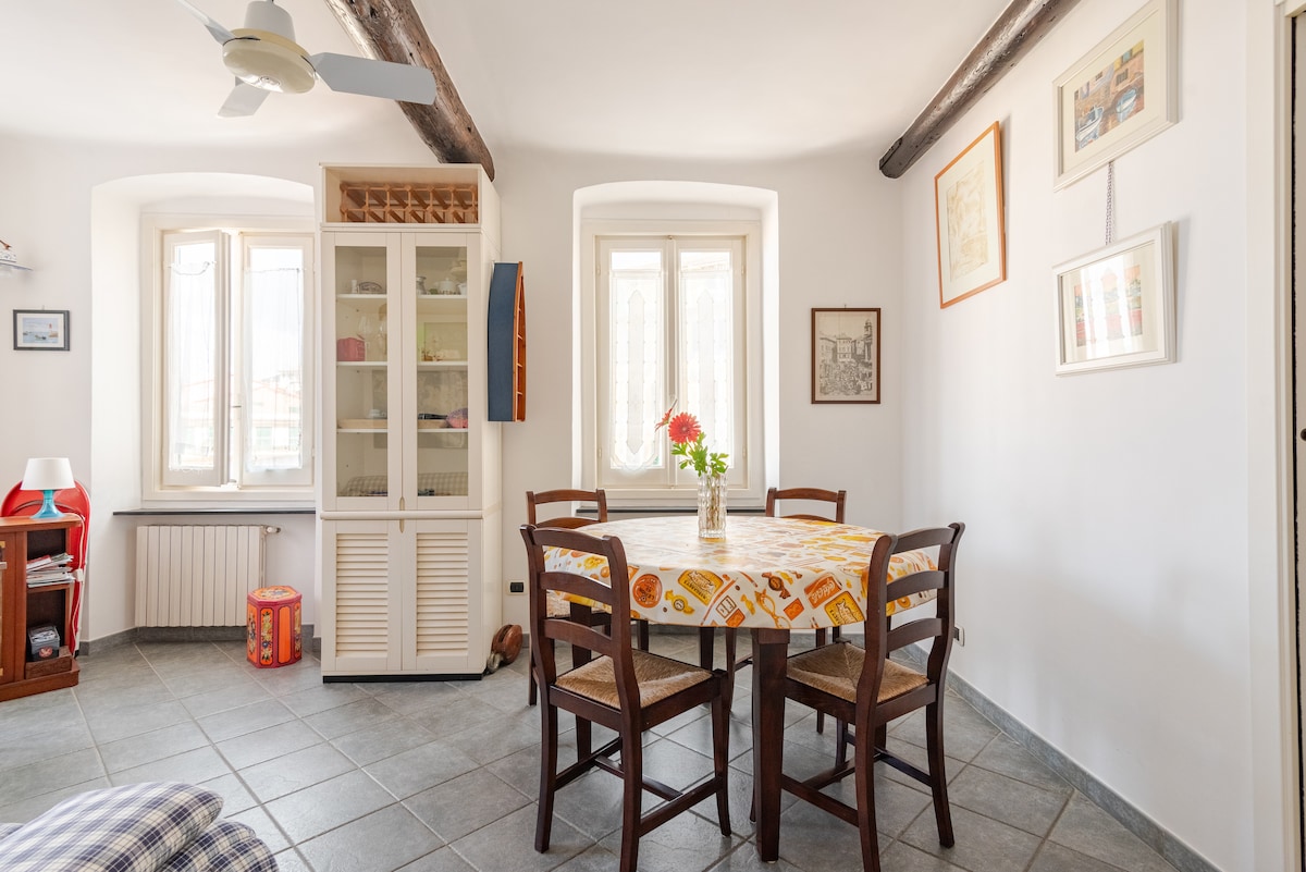 A bright dining area features a round table adorned with a colorful tablecloth and surrounded by four wooden chairs. Large windows allow ample natural light, and a glass-fronted cabinet adds storage space. Neutral tones and tiled flooring contribute to the room's open feel.