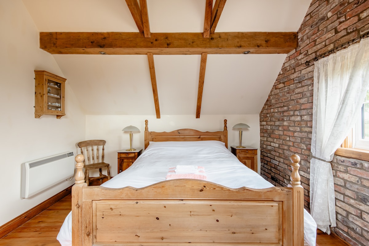 A spacious bedroom features a wooden bed frame with a light-colored mattress. The room showcases exposed wooden beams and warm wooden flooring. Flanking the bed are bedside tables with lamps, and a wooden chair is positioned nearby. A large window allows natural light to fill the space.
