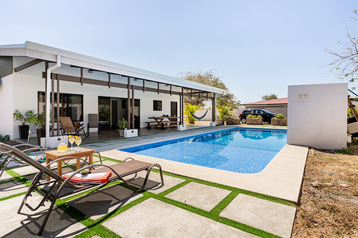 An outdoor space features a shimmering swimming pool surrounded by lounge chairs. A small table with drinks sits beside the pool, while a covered terrace offers seating for dining. The house is positioned in the background, framed by tropical greenery.