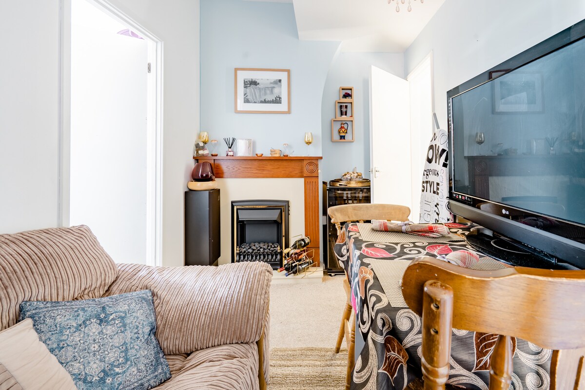 A cozy lounge area features a striped sofa complemented by a decorative cushion. An elegant wooden dining table is set with a tablecloth, while a television is positioned near a traditional fireplace. Bright light enters through an adjacent door, enhancing the space.