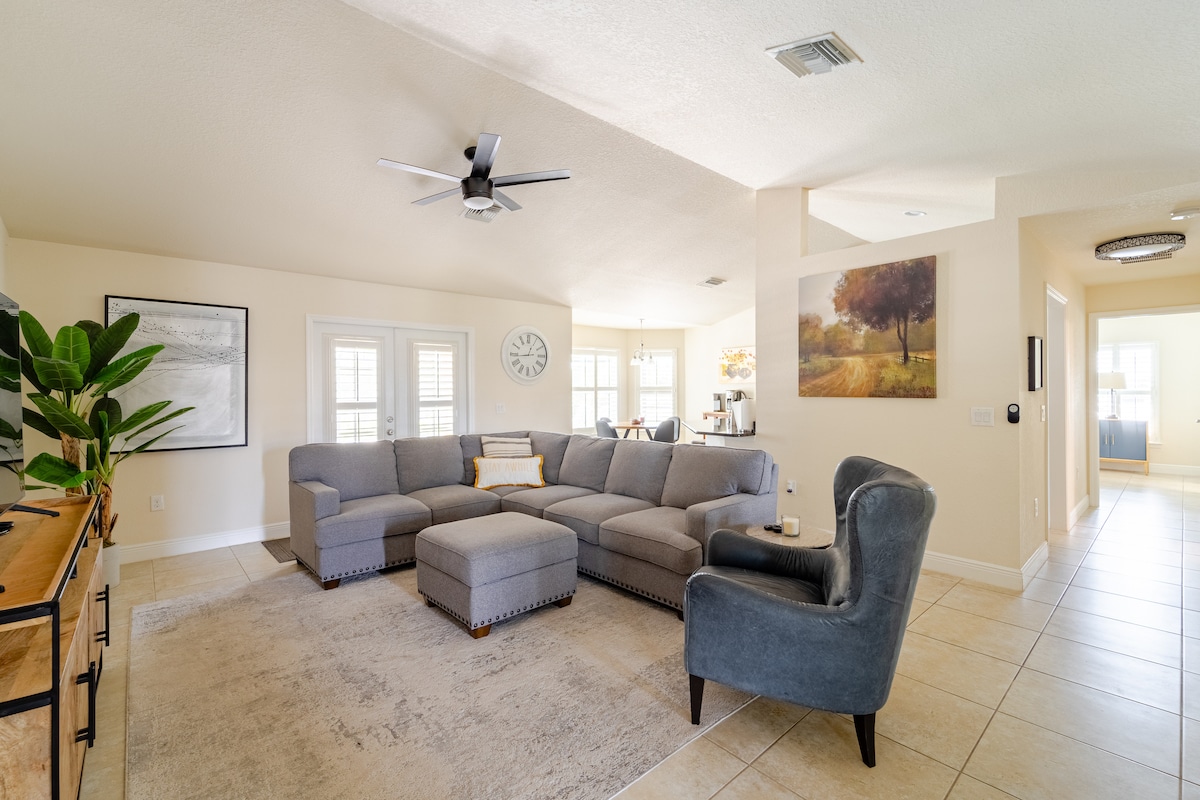 A spacious living room is presented with a large sectional sofa, accompanied by a single armchair. Natural light filters through the windows, illuminating the neutral decor. A decorative plant stands in one corner, while artwork adorns the walls, adding a touch of elegance.