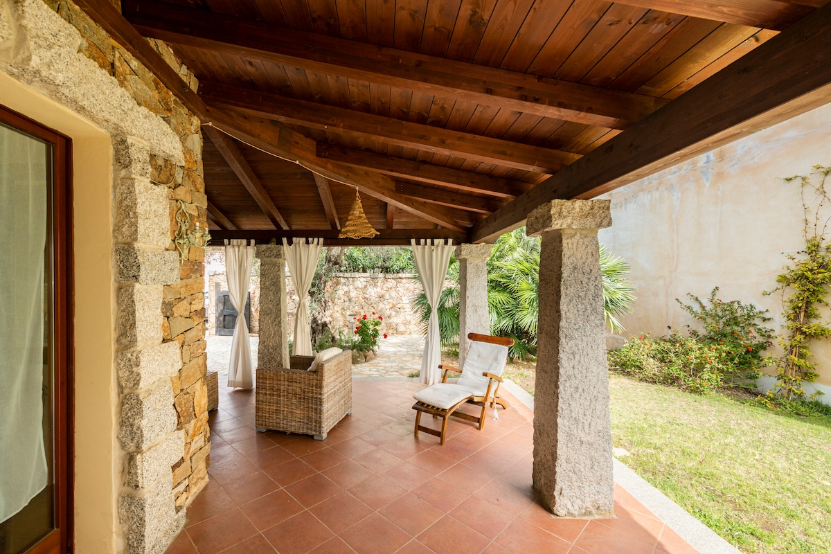 A shaded terrace is presented, featuring wooden beams and stone columns. Comfortable seating arrangements are visible, alongside soft drapes gently framing the space. Lush greenery and flowering plants add a natural touch to the inviting outdoor area, providing a serene environment.