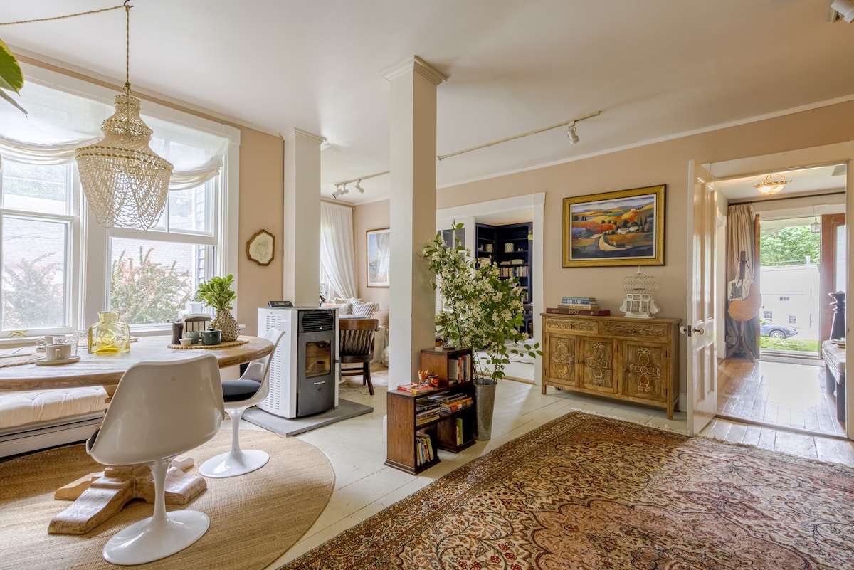 A bright, open living area features a round dining table with sleek white chairs and a cozy stove nearby. A decorative rug adds warmth to the space. Large windows allow natural light to fill the room, with a view of greenery outside.