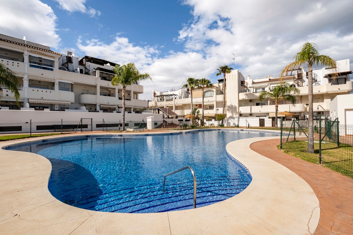 A large swimming pool is situated within a well-maintained residential complex, surrounded by palm trees and manicured grass areas. The pool features a curvy shape with a gentle entry point. Residential buildings are visible in the background, contributing to a serene atmosphere.