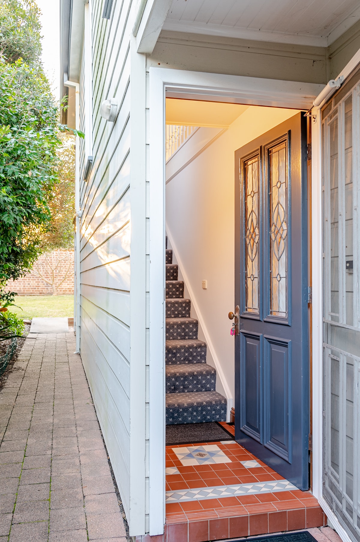 An inviting entrance is highlighted by a blue front door, leading to a staircase lined with patterned carpet. The warm lighting casts a welcoming glow over the tiled floor and surrounding exterior, framed by greenery from the adjacent garden.