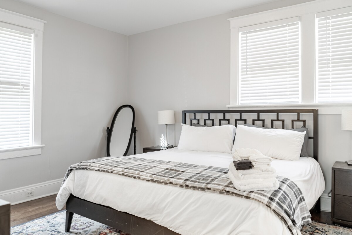 这间宽敞的一楼卧室配有一张加大双人床，配有清爽的白色床上用品和时尚的几何床头板。 柔软的灰色墙壁和大窗户营造出通风的感觉。 带灯的床头柜增加了温暖，而格子投掷则增强了舒适和时尚。