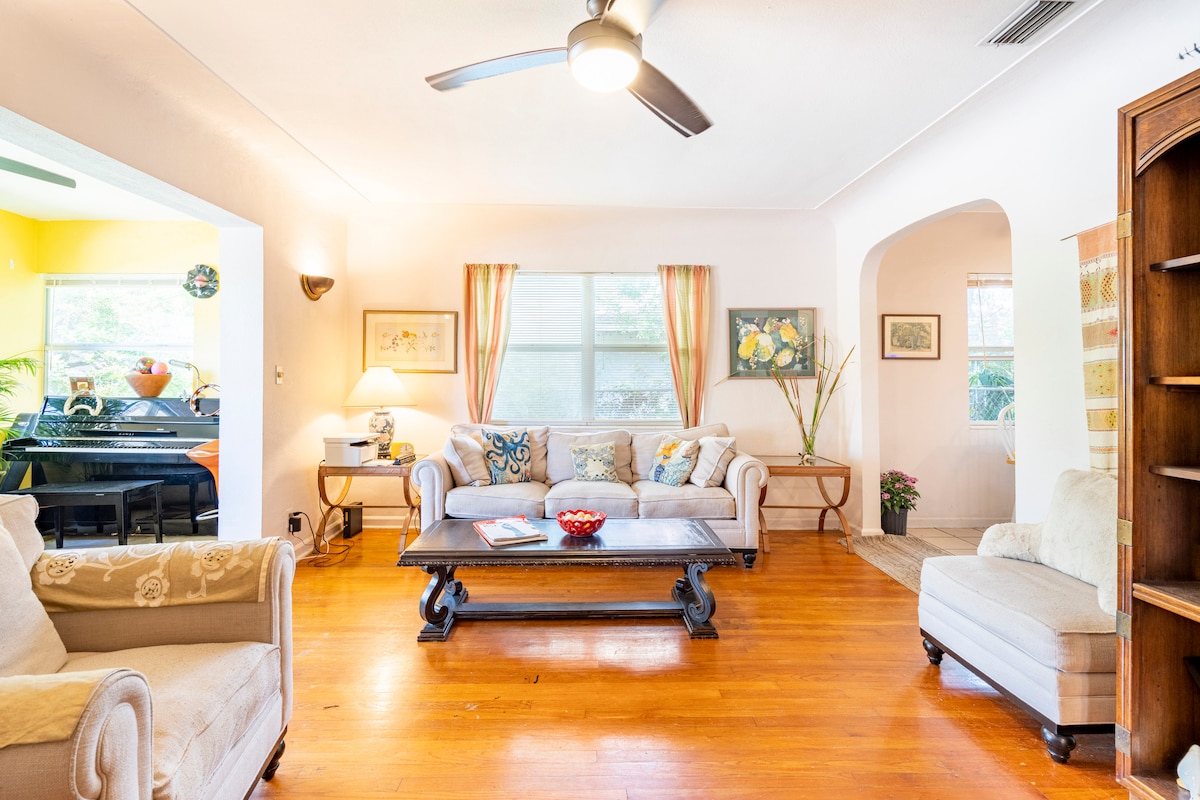 A bright living room features natural wood flooring, complemented by light curtains and a comfortable sofa. A coffee table occupies the center space, while a decorative cabinet and potted plant enhance the room's inviting atmosphere. Light streams in through the large windows.