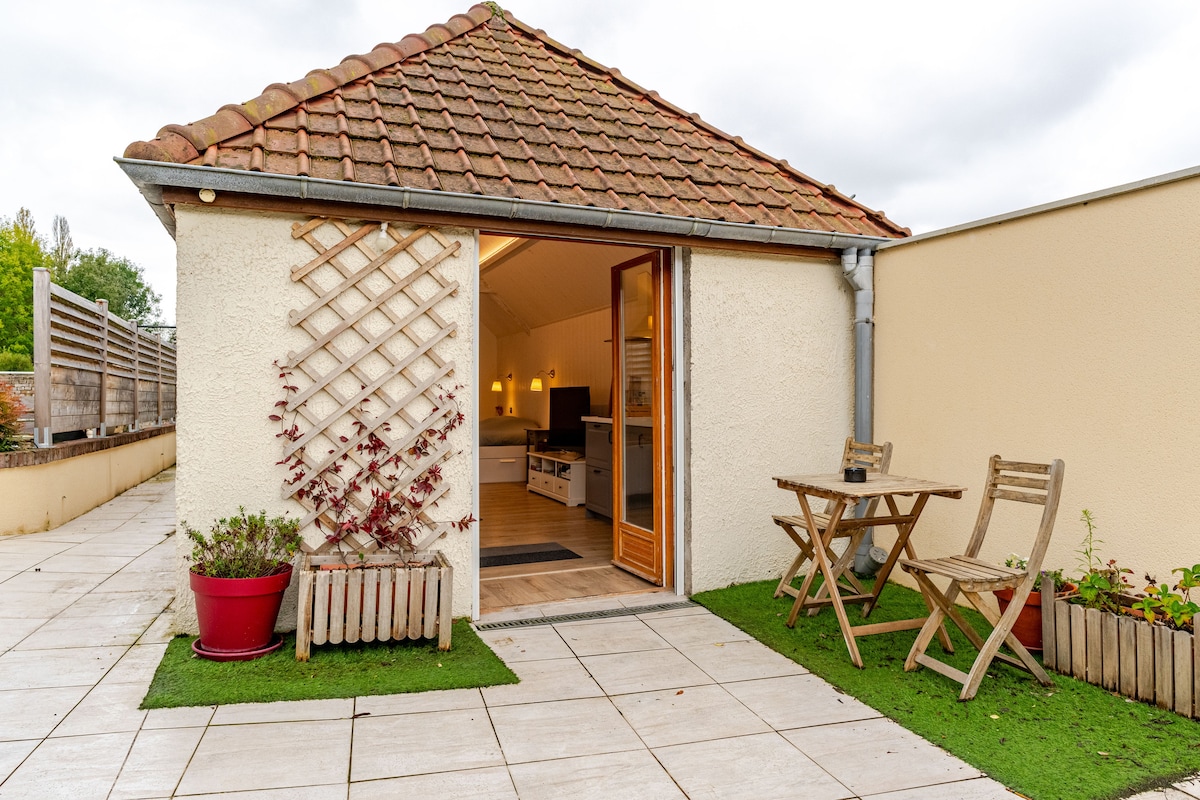 An outdoor patio area features a small table and two chairs, inviting relaxation. Surrounding greenery and flower beds enhance the space. The entrance to the studio is framed by decorative trellising, with a tiled pathway leading to the door.